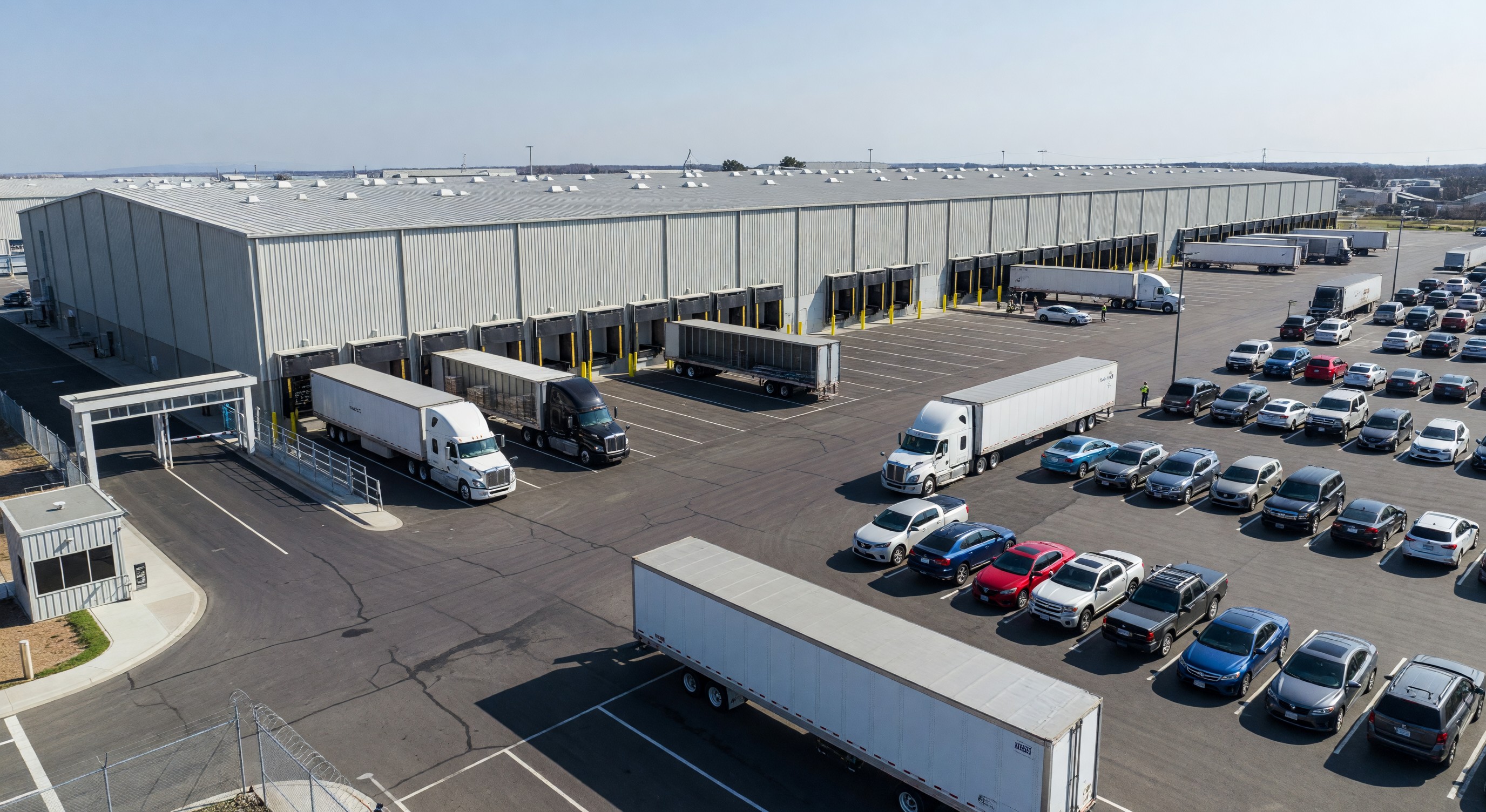 Large distribution center with truck staging yard, employee parking lot, and access control gate at entrance